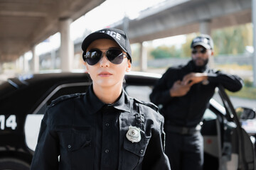 Wall Mural - confident policewoman in sunglasses looking at camera with blurred african american colleague on background outdoors.