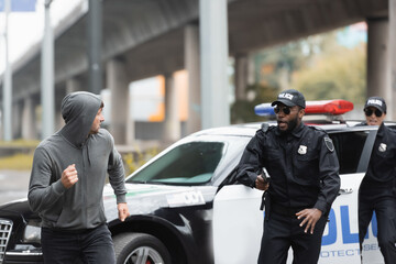 Wall Mural - Hooded offender running from multicultural police officers near patrol car on blurred background on urban street.