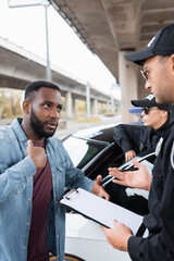 Wall Mural - African american victim talking with policeman holding clipboard on blurred background outdoors.