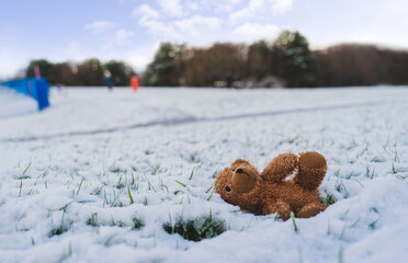 Wall Mural - Lost Teddy bear with sad face lying on snow with blurry people,Lonely bear doll laying down on the playground in winter, Lost toy or Loneliness concept, International missing Children