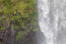 Young Man In A Helmet And Safety Harness Climbing A Mossy Cliff With A Rope