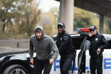 Poster - hooded offender running from shocked multicultural police officers on blurred background on urban street.