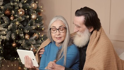 Wall Mural - Front view of senior couple indoors at home at Christmas, having video call with family. Elderle woman and man wrapped in blanket sit on sofa in cozy decorated room.