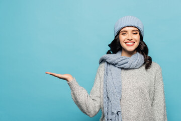 Front view of woman in winter outfit pointing with hand while looking at camera isolated on blue