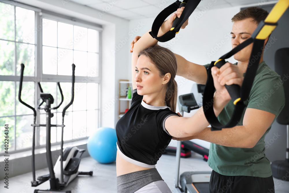 Coach helping young woman to train with TRX straps in gym