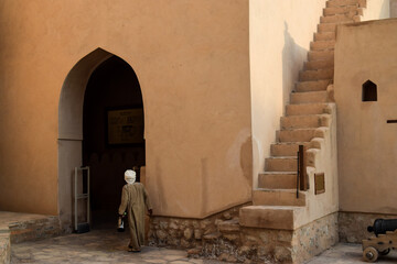 Back view of a man wearing traditional omani clothes walking inside Nizwa Fort next to a staircase and a huge door in the shape of an arch. Nizwa, Oman.