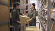 two warehouse asian chinese female worker colleague passing cardboard box in large depot of factory. lady giving parcel to coworker in stockroom from hand truck. storehouse woman staffs in teamwork