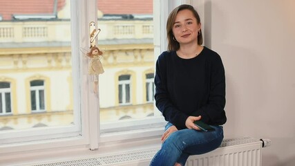 Poster - A woman sits near a window using a phone in her hands, checking notifications, looking at the camera, posing. Spending free time during a break in work. Waiting for a meeting.