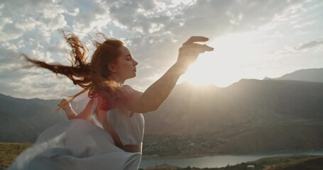 Poster - Woman in white dress standing on top of a mountain with raised hands while wind is blowing her dress and red hair - freedom, nature concept 4k footage