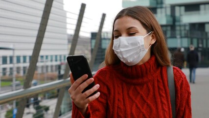 Wall Mural - Funny cheerful young woman with protective mask having a video chat talking smiling uses smartphone in city street. Happy cheerful girl video calling outdoors.