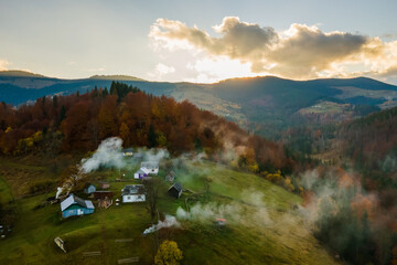 Aerial view of distant village with small shepherd houses on wide hill meadows between autumn forest trees in Ukrainian Carpathian mountains at sunset.