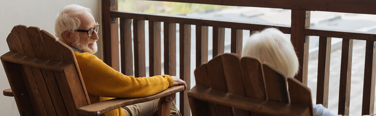 smiling senior husband looking at wife in armchair on terrace on blurred background, banner