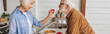 © LIGHTFIELD STUDIOS - side view of happy elderly wife feeding husband with cherry tomato in kitchen, banner