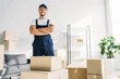 © LIGHTFIELD STUDIOS - cheerful mover in uniform and cap standing with crossed arms near boxes in apartment