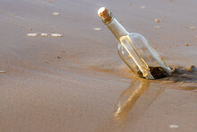 A Message In A Corked Bottle Washed Up On A Beach. The Bottle Is Reflected In The Wet Sand Below. The Bottle Is Standing At An Angle. Sunny Day.