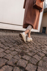 Wall Mural - Rear view on modern young woman in long elegant coat in beige pants in leather stylish youth shoes. Fashionable girl in spring casual clothes walks along stone road in city. Casual fashion .Close-up