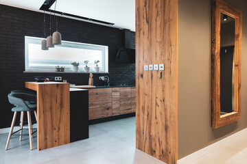 Wall Mural - Black brick wall in elegant kitchen with wooden island and stylish chairs, mirror in wooden frame on the wall