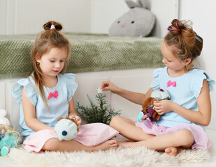 Two kid girls sisters in home dresses spend time together play with their favorite dolls at home look and talk. Happy childhood, cheerful lifestyle, games, comfortable pastime, hobby concept