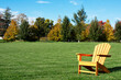 Traditional curveback sunset plastic outdoor patio adirondack chair with contoured backs and seats on green grass of manicured lawn surrouned by autumn trees.