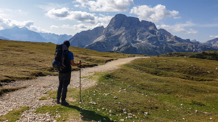 a man with hiking backpack hiking along a gravelled road in italian dolomites. there is a massive mo
