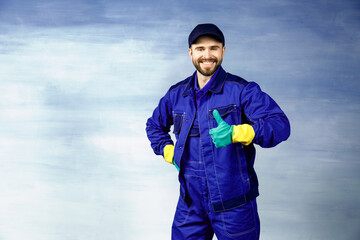  Thumb up. Young handsome man with a beard in a blue working uniform for cleaning rooms smiles isolated on blue background