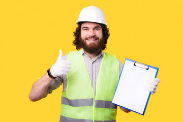 Wall Mural - Positive young bearded engineer is holding his work . pappers and looking at the camera is showing a thumb up.