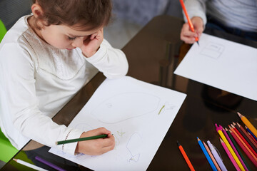 Top view of a baby girl in white shirt focused on drawing with color pencil. Color wooden pencils lying on a table.