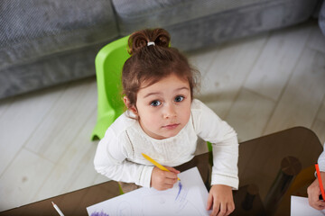 Top view of a cute preschool girl wearing white shirt and looking at camera. Adorable girl drawing indoors.