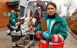 © Valerii Apetroaiei - A woman paramedic in uniform stands with a patient card in front of an ambulance and her colleague standing near a patient's gurney.
