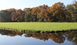 Autumn landscape with water in the foreground, a green pasture and trees in the back. The trees reflecting in the still water