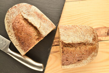 Two halves of a loaf, fragrant buckwheat bread with a metal knife and a serving board of slate, close-up, on a wooden table.