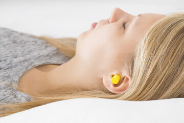 Wall Mural - Young adult blonde woman sleeping with yellow earplug into her ear in bed. Head on pillow. Very well asleep in silence. Side view. Closeup.