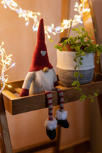 Christmas Elf Sitting On A Wooden Shelf Next To A Green Plant, Illuminated By Light Garlands. Cute Christmas Decoration.