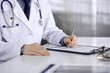 © rogerphoto - Unknown male doctor sitting and working with clipboard of medication history record in clinic at his working place, close-up. Young physician at work. Perfect medical service, medicine concept