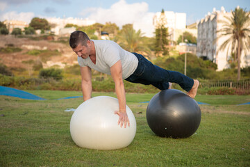 Fitness coach balancing himself on top of 2 fit-balls as part of an exercise. Skating pump track in the background.