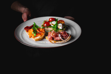 chef in a black suit holds in his hands plate with Bruschetta on Dark grey black background