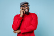 Cheerful handsome young bearded african american man 20s in casual red shirt cap eyeglasses standing put hand prop up on chin looking camera isolated on pastel blue color background studio portrait.