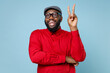 Cheerful funny young bearded african american man 20s wearing casual red shirt cap eyeglasses standing showing victory sign looking camera isolated on pastel blue color background studio portrait.