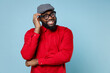 Confused pensive puzzled young bearded african american man 20s wearing casual red shirt eyeglasses cap standing put hand on head looking camera isolated on pastel blue background studio portrait.