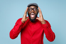 Surprised Excited Young Bearded African American Man In Casual Red Shirt Cap Eyeglasses Standing Screaming With Hands Gesture Near Mouth Looking Camera Isolated On Blue Background Studio Portrait.