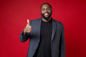 Smiling handsome confident young african american business man 20s wearing classic jacket suit standing showing thumb up looking camera isolated on bright red color wall background studio portrait.