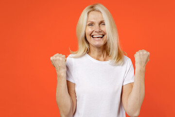 Wall Mural - Funny joyful elderly gray-haired blonde woman lady 40s 50s years old in white casual t-shirt standing doing winner gesture clenching fists isolated on bright orange color background studio portrait.