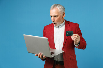 Shocked elderly gray-haired mustache bearded business man wearing red jacket suit standing working on laptop pc computer hold credit bank card isolated on blue color wall background studio portrait.