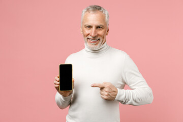 Smiling elderly gray-haired mustache bearded man in casual white turtleneck standing point index finger on mobile cell phone with blank empty screen isolated on pastel pink background studio portrait.