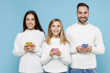 Wall Mural - Cheerful young parents mom dad with child kid daughter teen girl in white sweaters using mobile phone typing sms message isolated on blue background studio. Family day parenthood childhood concept.