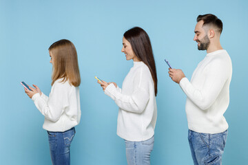 Wall Mural - Side view of smiling young parents mom dad with child kid daughter teen girl in white sweaters using mobile phone typing sms message isolated on blue background studio. Family day parenthood concept.