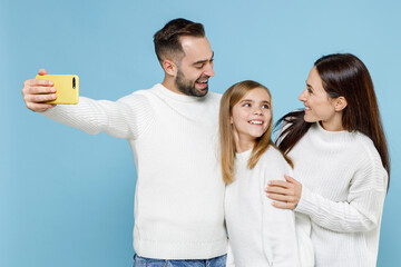 Wall Mural - Pretty young parents mom dad with child kid daughter teen girl in white sweaters doing selfie shot on mobile phone hugging isolated on blue background studio. Family day parenthood childhood concept.