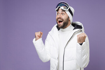 Wall Mural - Excited skier man in warm white windbreaker jacket ski goggles mask doing winner gesture spend extreme weekend winter season in mountains isolated on purple background. People lifestyle hobby concept.