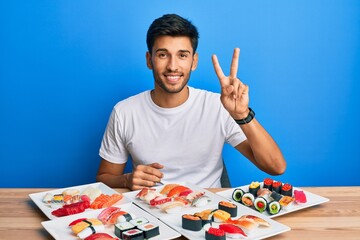 Young handsome man eating sushi sitting on the table showing and pointing up with fingers number two while smiling confident and happy.