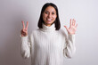 © Roquillo - Young brunette woman wearing white knitted sweater against white background showing and pointing up with fingers number seven while smiling confident and happy.